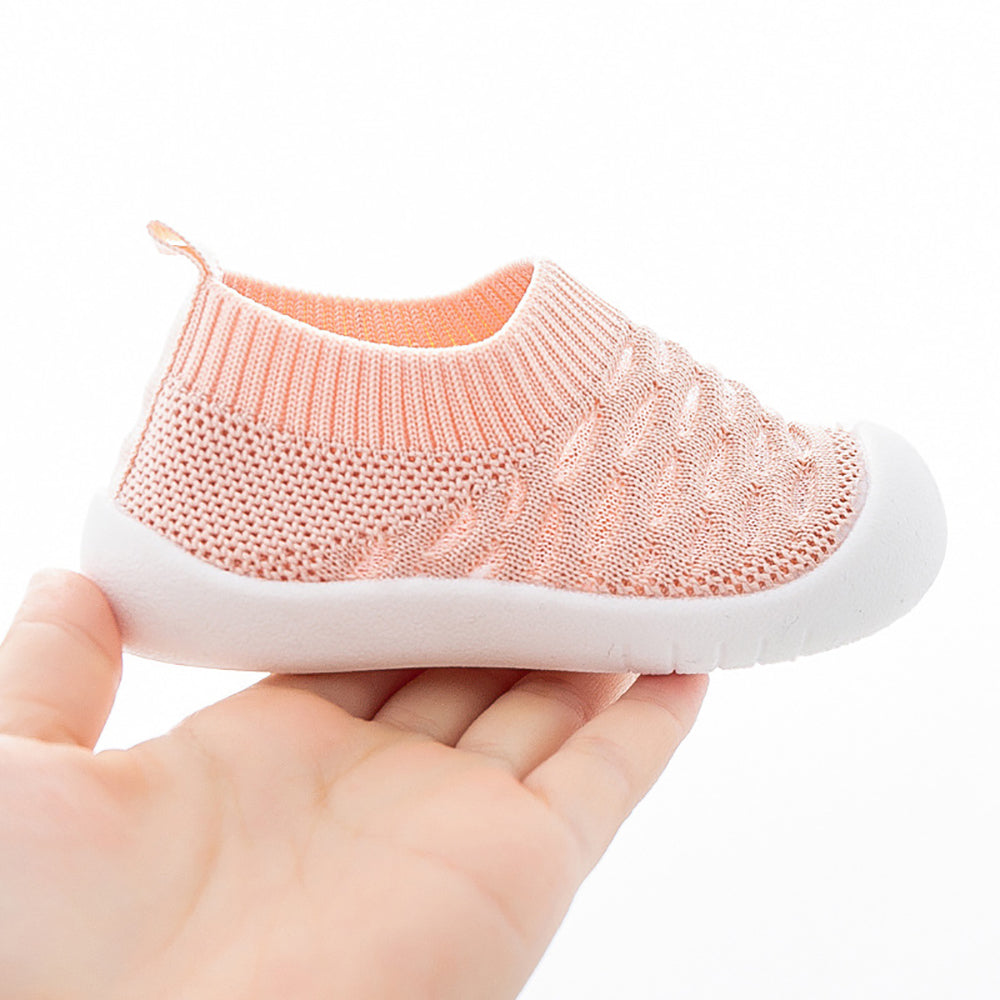 Hand holding coral breathable baby shoe showing knit texture and slip-resistant sole